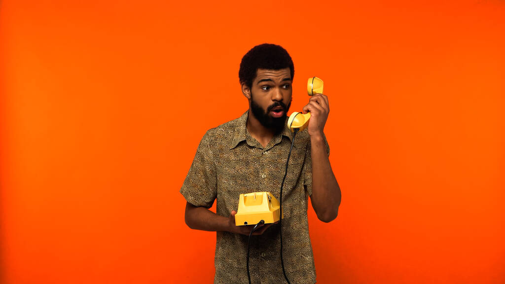 σοκαρισμένος Αφροαμερικανός με γενειάδα κρατώντας κίτρινο ρετρό τηλέφωνο σε πορτοκαλί φόντο  - Φωτογραφία, εικόνα