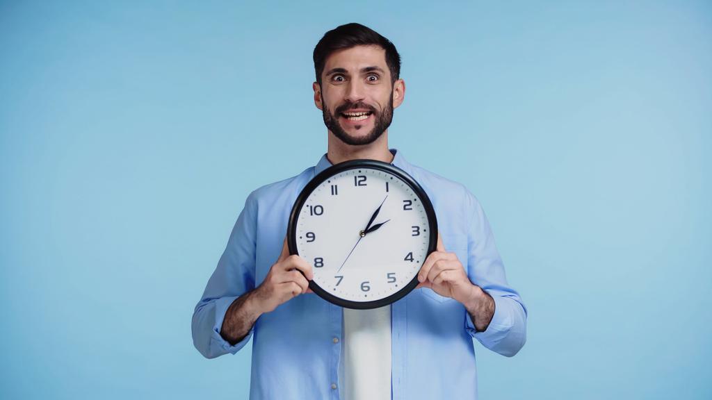 приголомшений чоловік у сорочці тримає годинник, дивлячись на камеру ізольовано на синьому фоні
  - Фото, зображення