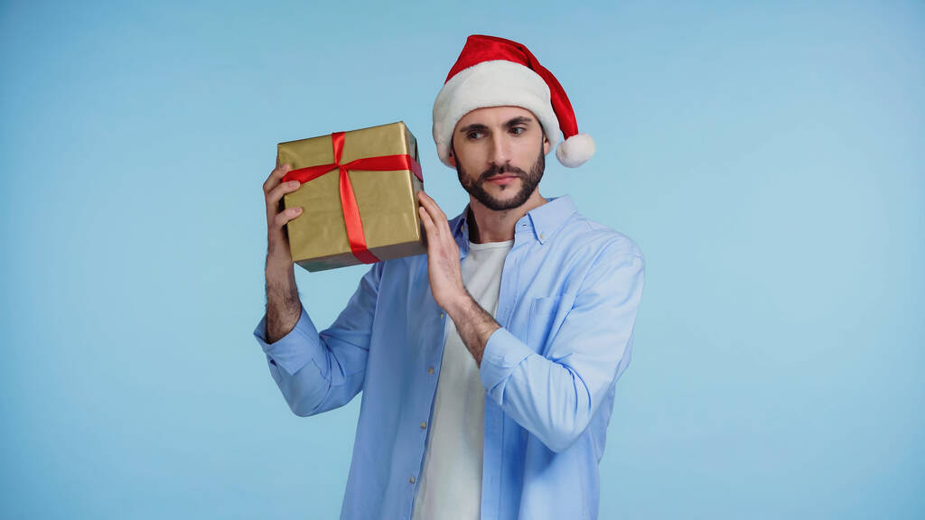 Kırmızı Noel Baba şapkalı, tuhaf bir adam mavi renkte Noel hediyesini sallıyor. - Fotoğraf, Görsel