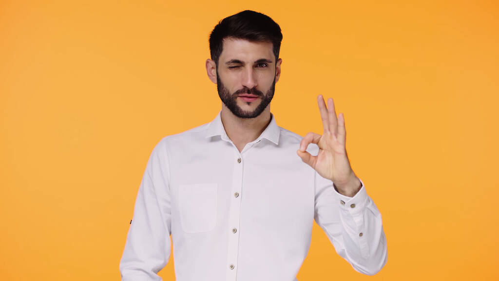 γενειοφόρος άνδρας σε λευκό πουκάμισο δείχνει ok σημάδι, ενώ κλείνει το μάτι απομονώνονται σε κίτρινο - Φωτογραφία, εικόνα