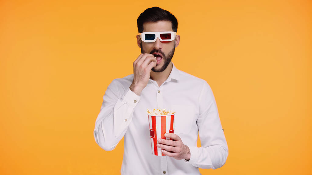 hombre barbudo en gafas 3d sosteniendo cubo rayado y comer palomitas de maíz aislado en amarillo - Foto, Imagen