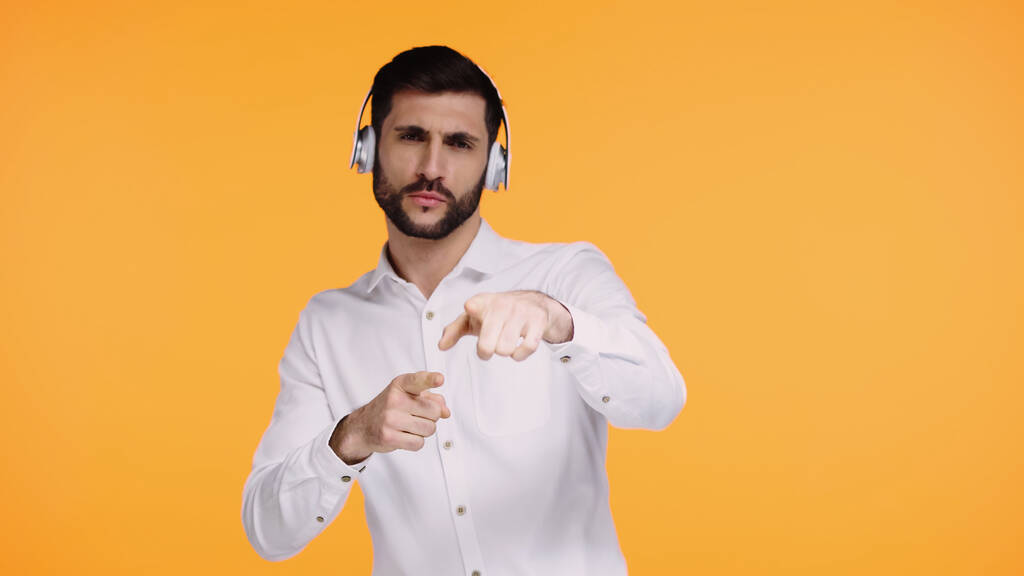 hombre barbudo en auriculares inalámbricos apuntando con los dedos y mirando a la cámara aislada en amarillo - Foto, imagen
