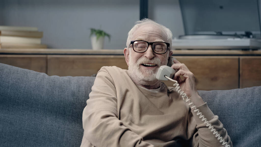 Χαρούμενος ηλικιωμένος άνδρας με γυαλιά ηλίου που μιλάει στο παλιό τηλέφωνο και κάθεται στον καναπέ στο σπίτι - Φωτογραφία, εικόνα