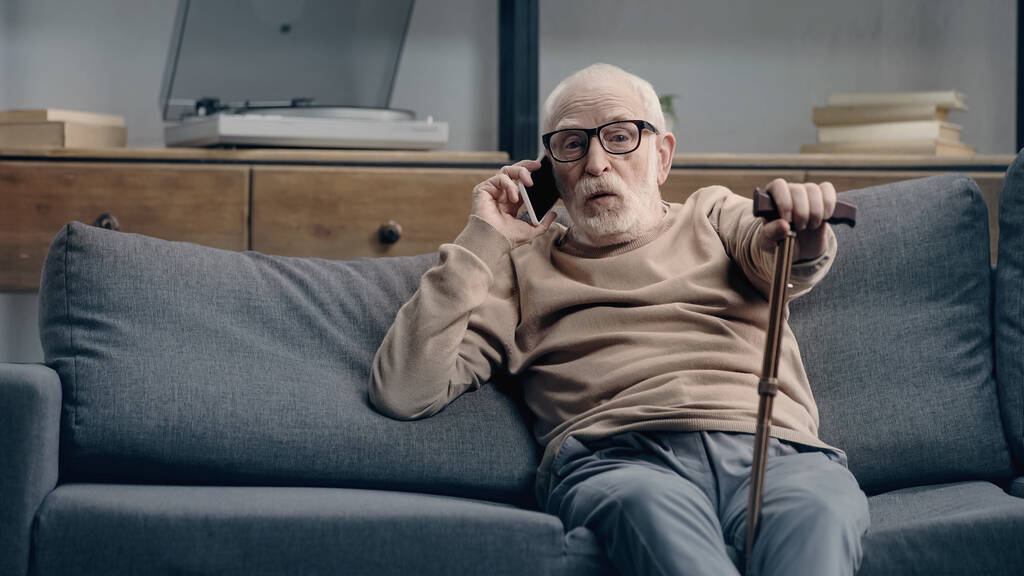 έκπληκτος ηλικιωμένος μιλάει στο smartphone και κάθεται στον καναπέ με το μπαστούνι  - Φωτογραφία, εικόνα