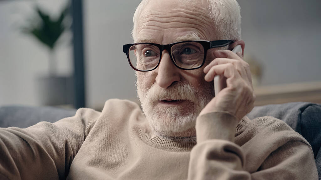 бородатый мужчина в очках разговаривает по мобильному телефону дома  - Фото, изображение