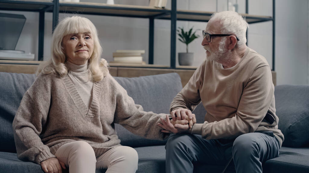 Старший мужчина успокаивает неуверенную жену со слабоумием, сидящую на диване - Фото, изображение