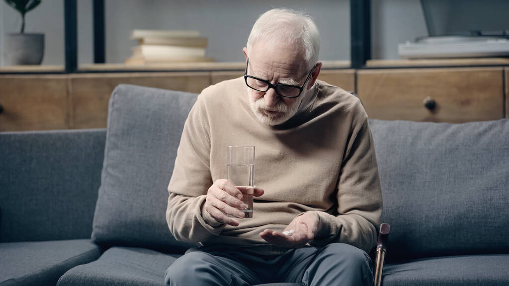 Idősebb férfi demenciával, aki tablettákat és egy pohár vizet tart otthon. - Fotó, kép