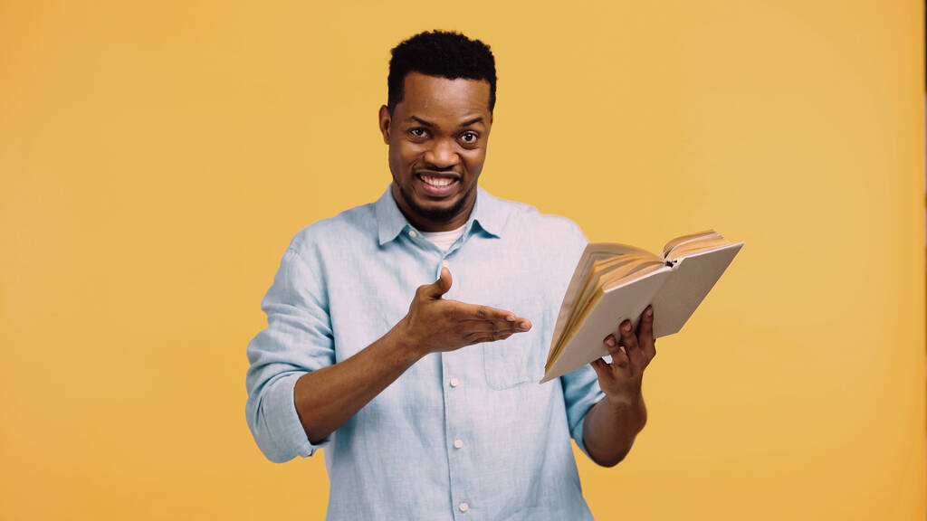 šťastný africký Američan v modré košili ukazuje rukou na knihu izolované na žluté - Fotografie, Obrázek