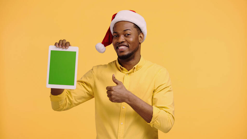 幸せなアフリカ系アメリカ人の男性サンタの帽子親指を表示し、黄色に隔離された緑の画面でデジタルタブレットを保持 - 写真・画像