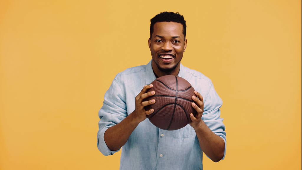 Χαρούμενος Αφροαμερικάνος που κρατάει το μπάσκετ απομονωμένο στο κίτρινο - Φωτογραφία, εικόνα