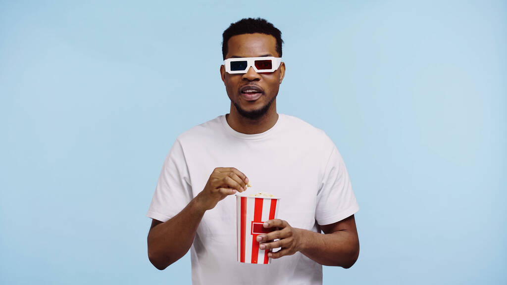 focused african american man in 3d glasses watching movie and holding popcorn isolated on blue - Photo, Image