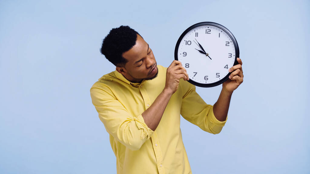 hombre afroamericano en camisa amarilla sosteniendo reloj aislado en azul - Foto, imagen