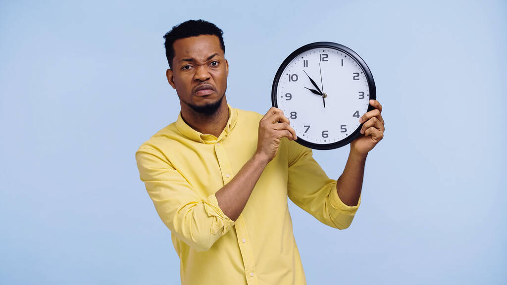 niezadowolony Afrykanin amerykański człowiek w żółtej koszuli trzymając zegar odizolowany na niebiesko - Zdjęcie, obraz