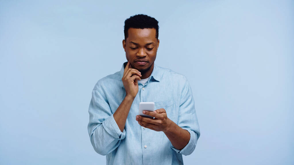 Pensive αφρικανός Αμερικανός με πουκάμισο χρησιμοποιώντας το κινητό τηλέφωνο απομονώνονται σε μπλε - Φωτογραφία, εικόνα