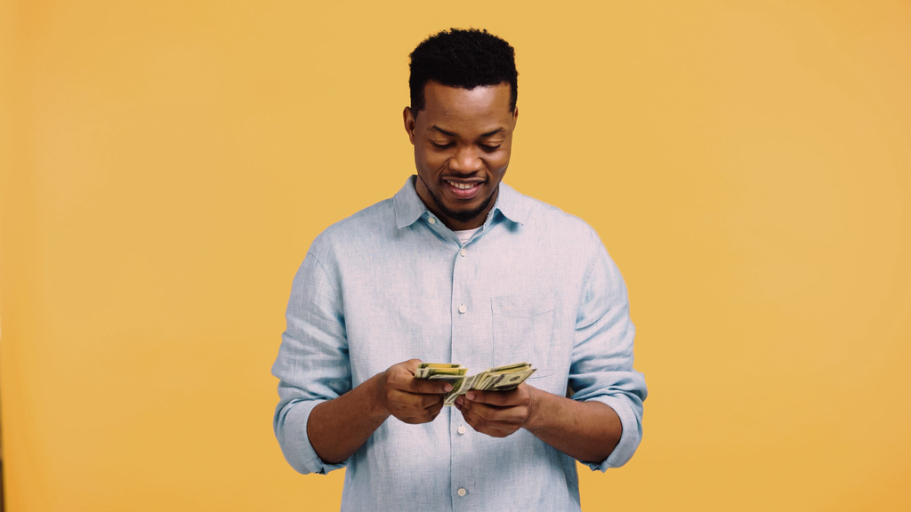 szczęśliwy Afrykanin amerykański człowiek w niebieskiej koszuli licząc banknoty dolarowe odizolowane na żółto - Zdjęcie, obraz