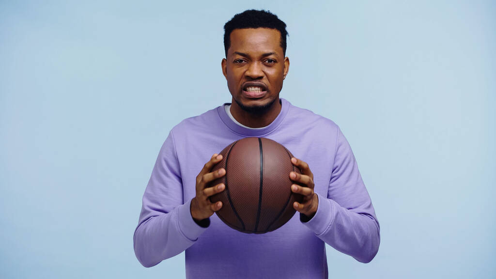 δυσαρεστημένος Αφροαμερικανός που κρατάει το μπάσκετ απομονωμένο στο μπλε.  - Φωτογραφία, εικόνα