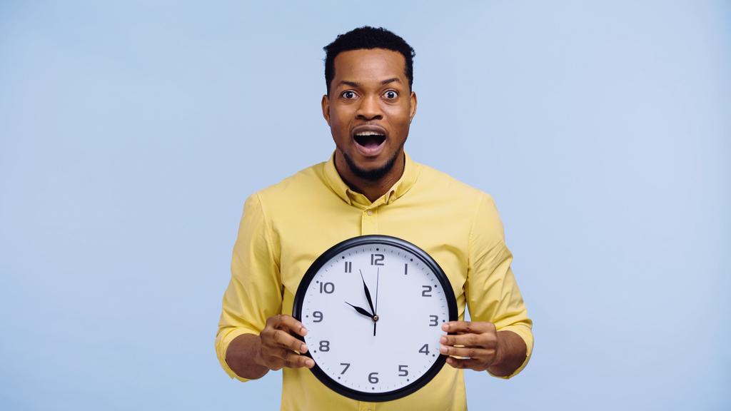 κατάπληκτος Αφροαμερικάνος που κρατάει το ρολόι και κοιτάζει την κάμερα απομονωμένη στο μπλε - Φωτογραφία, εικόνα