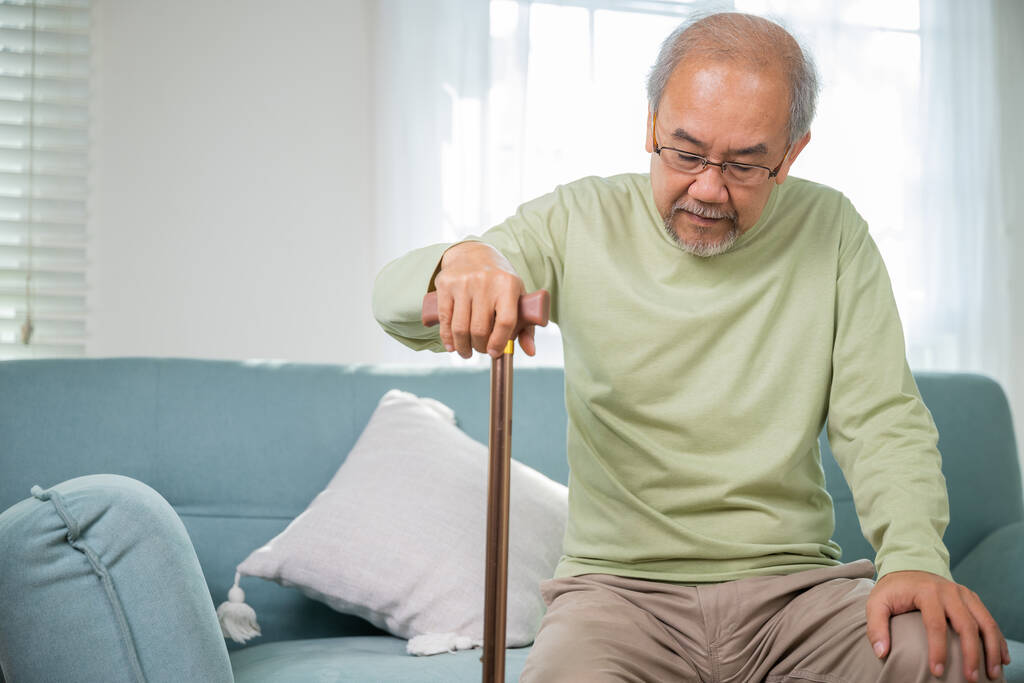 Ασιάτης γέρος με γυαλιά για να σηκωθεί από καναπέ με μπαστούνι, ηλικιωμένος που υποφέρει από πόνο στο γόνατο κρατώντας λαβή του ζαχαροκάλαμου, ηλικιωμένος με ειδικές ανάγκες κρατώντας μπαστούνι στο σπίτι - Φωτογραφία, εικόνα