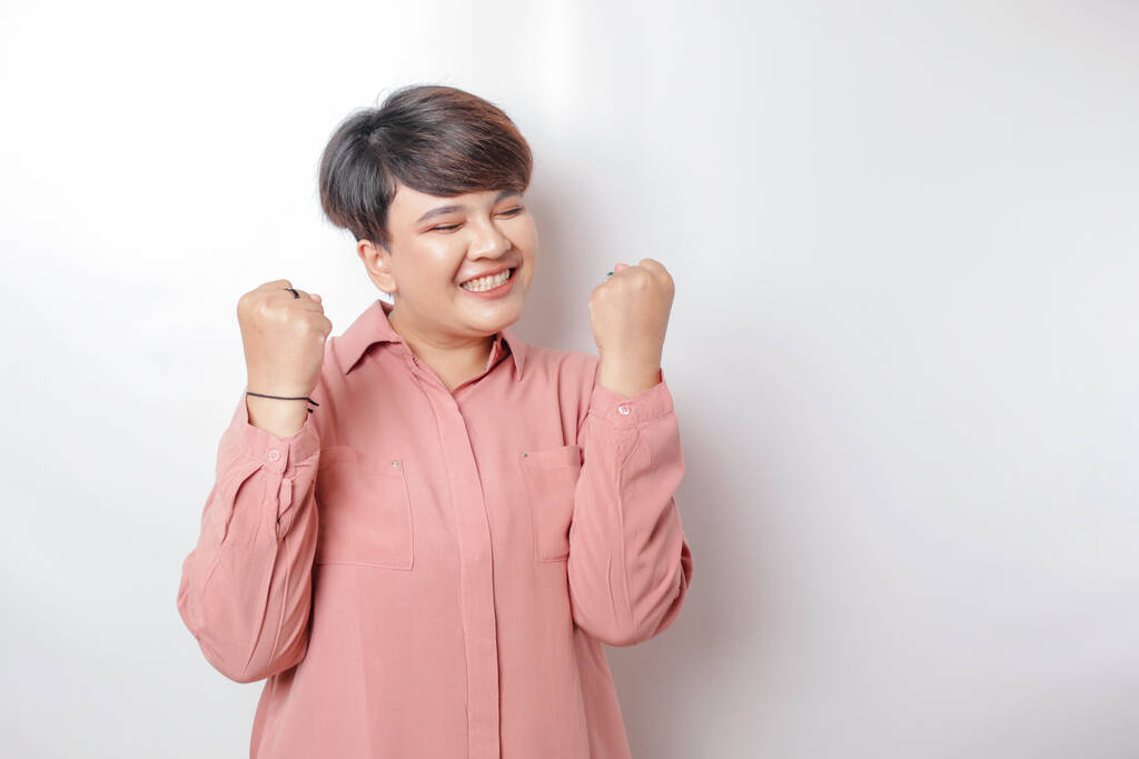 Eine junge, kurzhaarige Asiatin mit glücklichem, erfolgreichen Gesichtsausdruck trägt ein rosafarbenes Hemd vor weißem Hintergrund - Foto, Bild