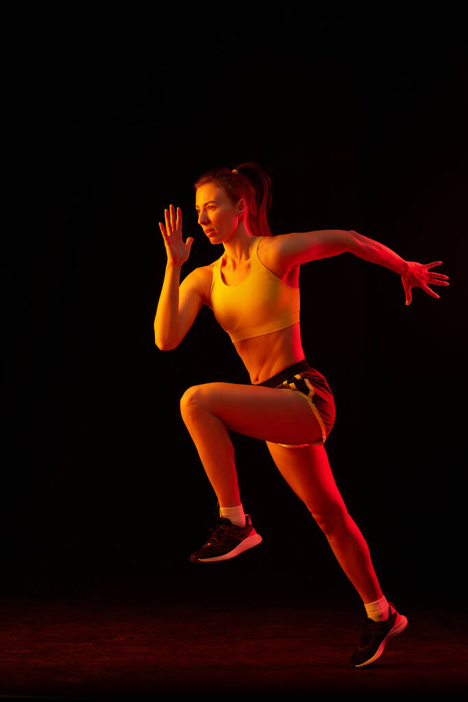 ネオンの光の中で暗い背景に隔離された実行している若いスポーツ女性のトレーニングの肖像画。競争、スポーツ、フィットネス、スピードとエネルギーの概念。ダイナミックな動き. - 写真・画像