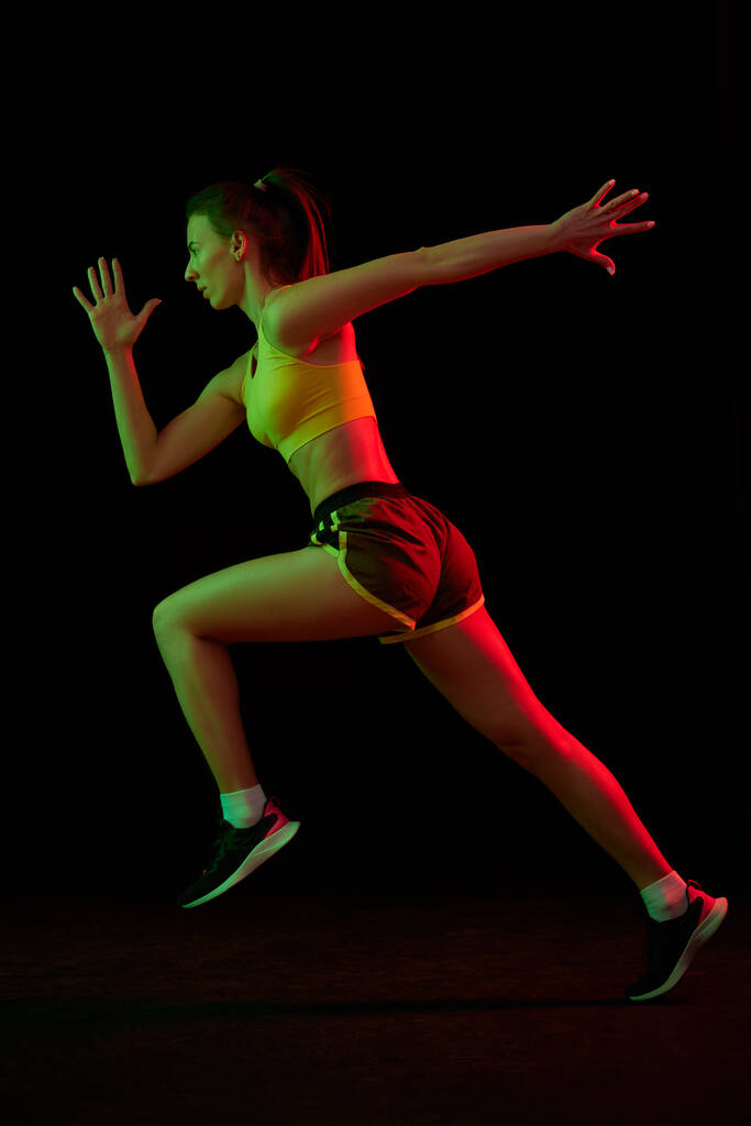 Běžící technika. Profesionální běžkyně běžící izolovaně na tmavém pozadí v neonovém světle. Zdravý životní styl, sport, fitness, rychlost a energie koncepce. Sportovní žena ukazující základní pohyby - Fotografie, Obrázek