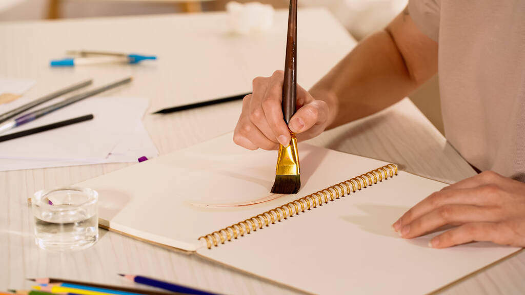 Обрезанный вид молодой женщины, рисующей на альбоме у воды и размытые цветные карандаши на столе  - Фото, изображение
