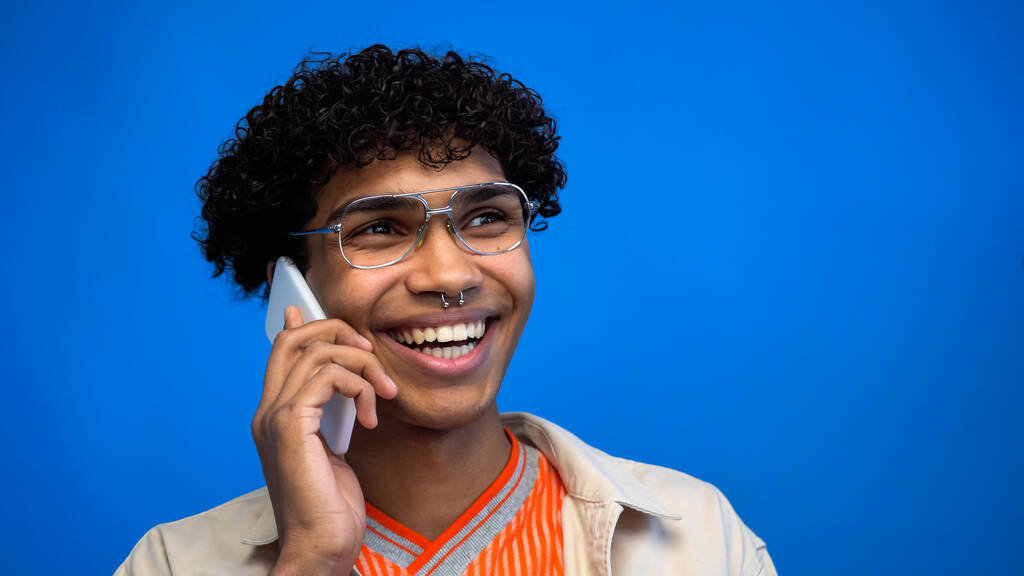 青で隔離された携帯電話で話している眼鏡の陽気なアフリカ系アメリカ人男性  - 写真・画像