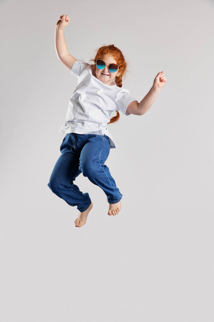 Heerlijk. Happy little girl, kid in white t-shirt en jeans springen hoog geïsoleerd over witte achtergrond. Kindermode, emoties, zorgeloze kindertijd, uitdagingen, onderwijsconcept - Foto, afbeelding