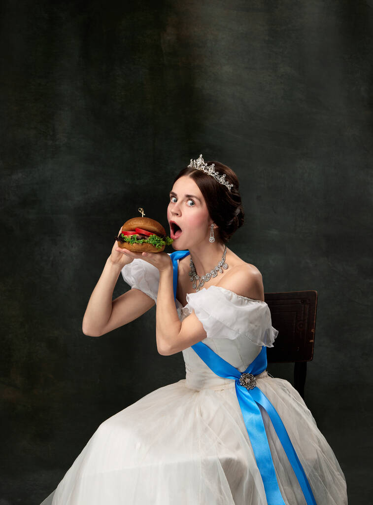 Bardzo smaczne. Emocjonalna kobieta, królewska osoba, królowa lub księżniczka w białym średniowiecznym stroju jedząca burgera na ciemnym tle. Koncepcja fast food, wyraz twarzy, porównanie ery, moda, piękno. - Zdjęcie, obraz