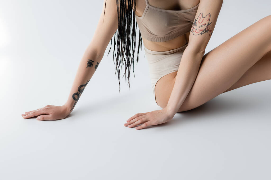 Oříznutý pohled na tetované ženy ve spodním prádle dotýkající se šedého pozadí  - Fotografie, Obrázek