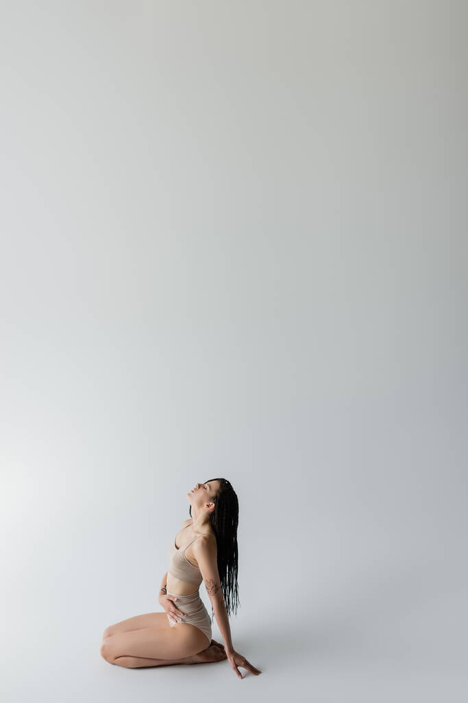 Zijaanzicht van jonge fitte vrouw in ondergoed zittend op grijze achtergrond  - Foto, afbeelding