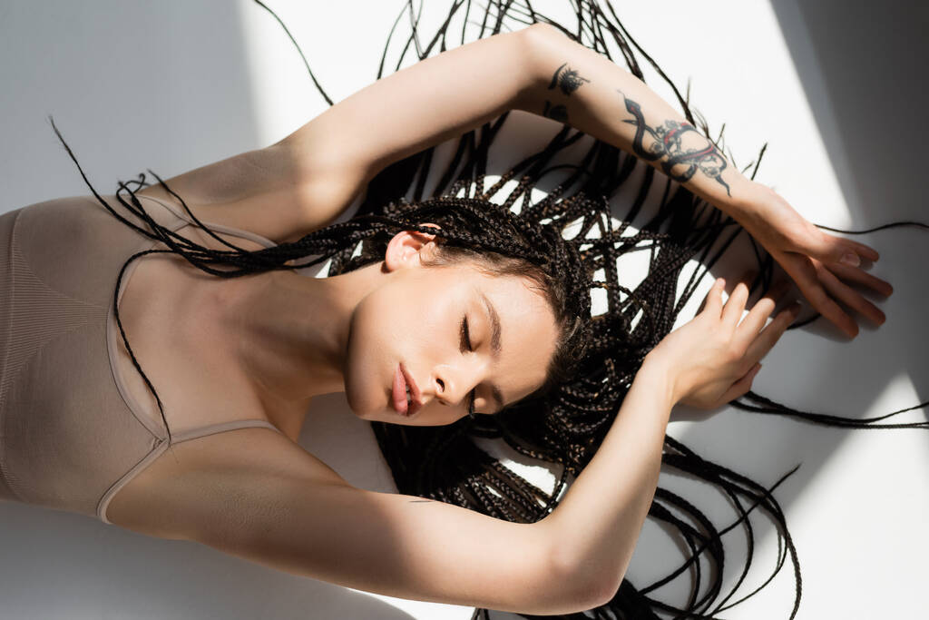 Visão superior da mulher tatuada com tranças deitadas sobre fundo cinza com luz  - Foto, Imagem