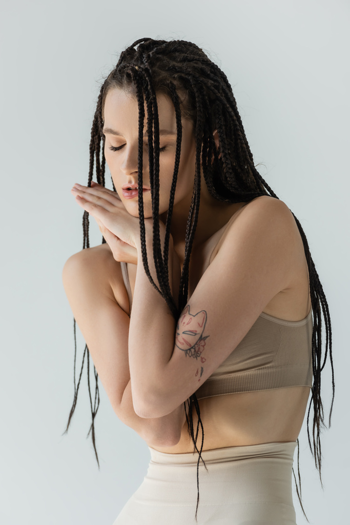Νεαρή γυναίκα με τατουάζ στο αθλητικό σουτιέν που κρατιέται χέρι-χέρι κοντά στο πρόσωπο απομονωμένη στο γκρι  - Φωτογραφία, εικόνα