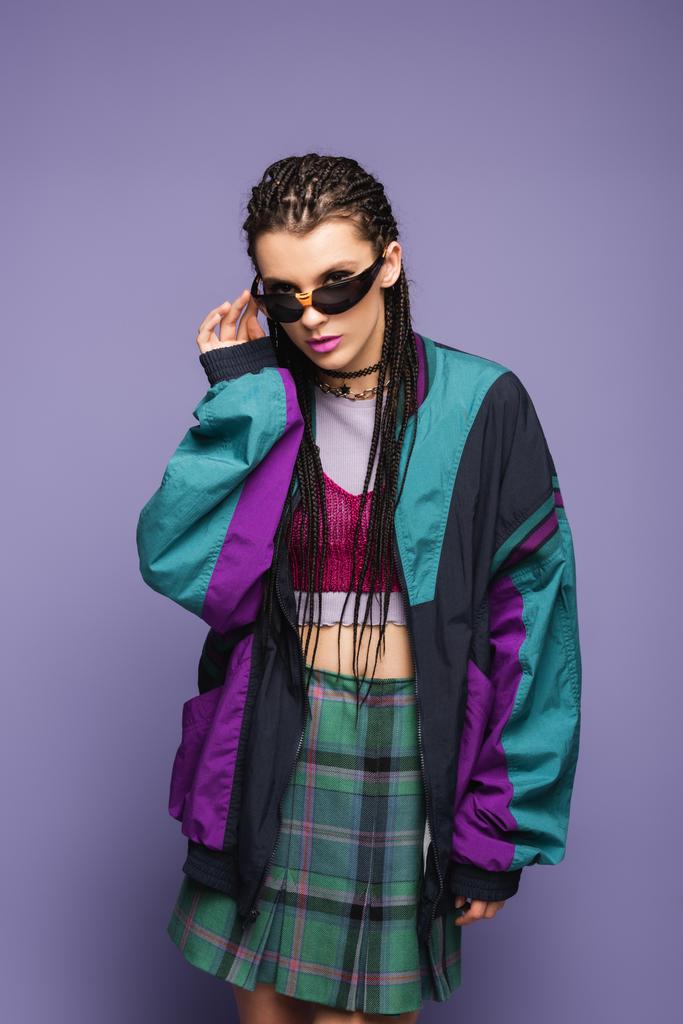 Young woman in retro sports jacket holding sunglasses on purple background  - Photo, Image