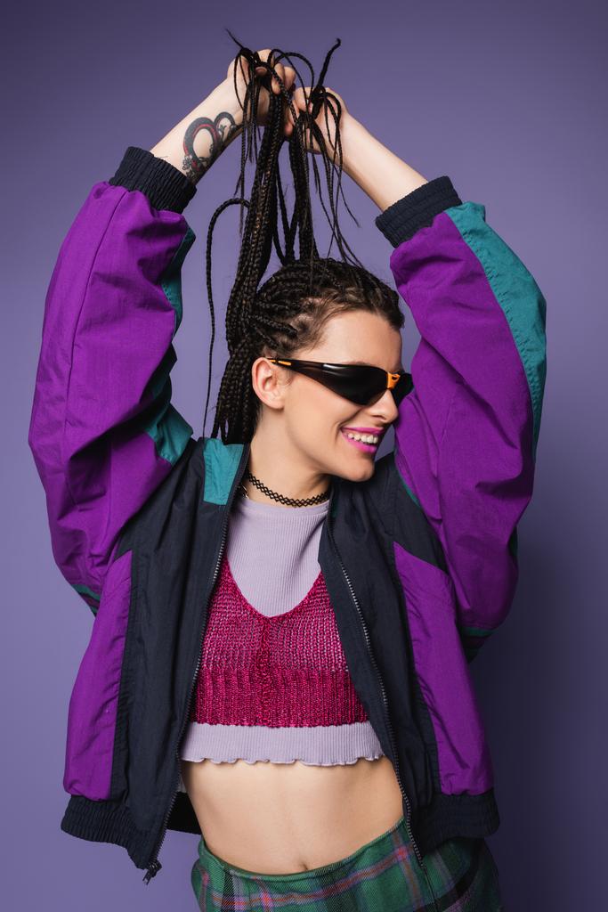 usmívající se žena v slunečních brýlích a vinobraní bunda drží copánky izolované na fialové - Fotografie, Obrázek