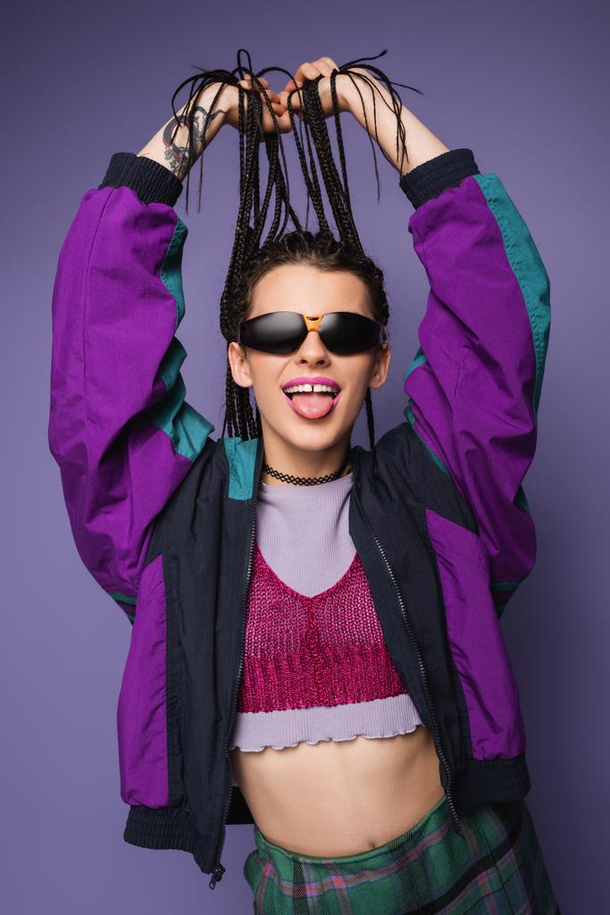 giovane donna in giacca vintage e occhiali da sole che tengono le trecce e sporgono la lingua isolata sul viola - Foto, immagini