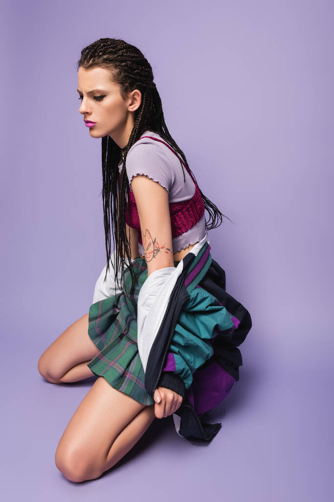 стильная татуированная женщина с плетеными дредами на коленях на фиолетовом фоне - Фото, изображение