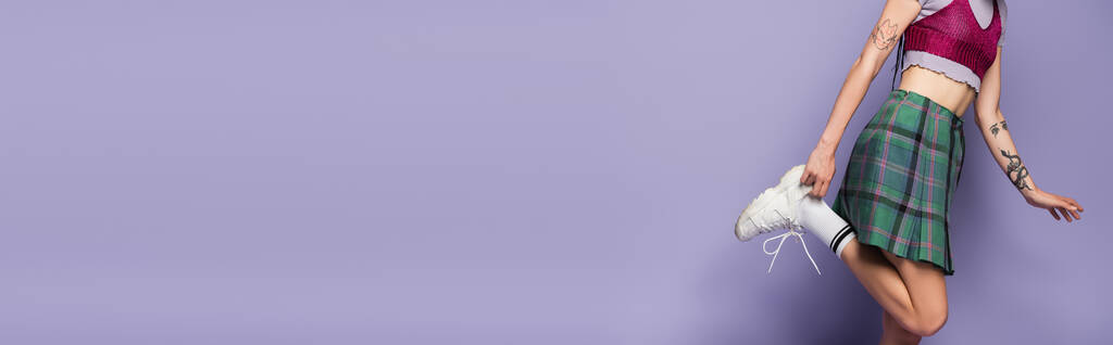 ostříhaný pohled na tetovanou ženu v kostkované sukni upravující bílou tenisku na purpurovém pozadí, banner - Fotografie, Obrázek