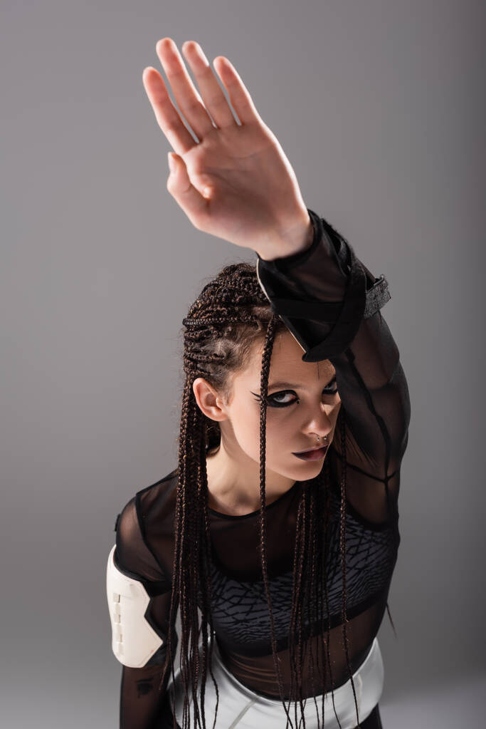 high angle view of woman with braids and futuristic style makeup and clothes posing with raised hand isolated on grey - Photo, Image
