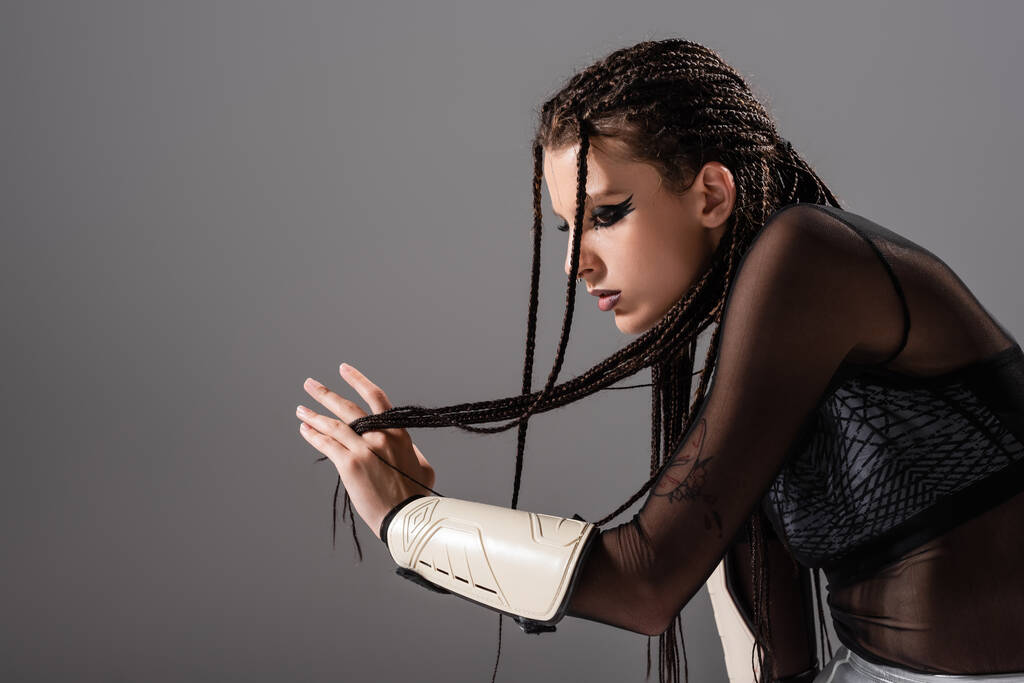 side view of futuristic woman in stylish makeup and outfit touching braided dreadlocks isolated on grey - Photo, Image
