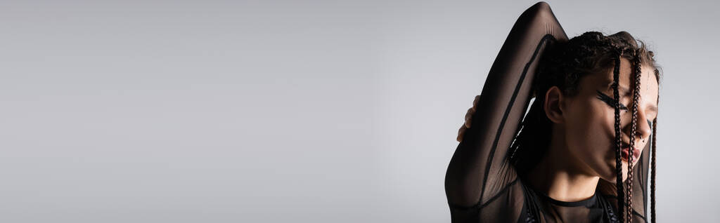brunetka žena s make-upem a copánky pózuje se zavřenýma očima a rukama za hlavou izolované na šedé, prapor - Fotografie, Obrázek