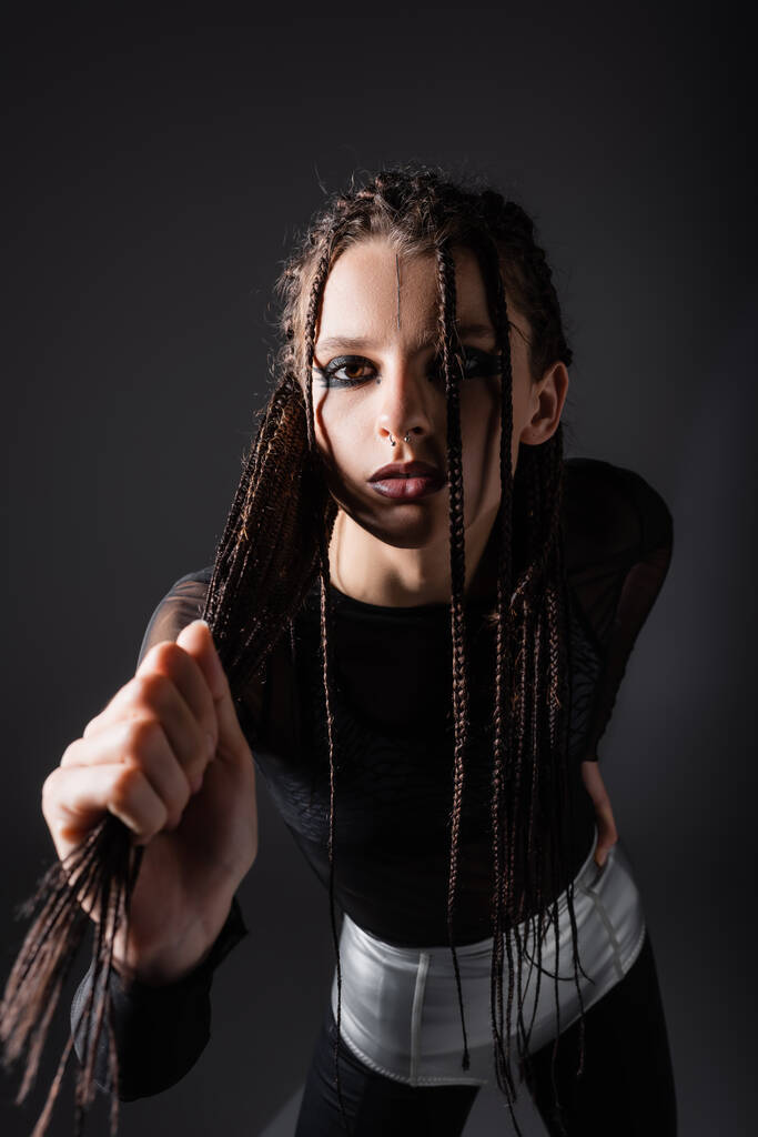 stylish woman with makeup holding braided dreadlocks and looking at camera isolated on black - Photo, Image