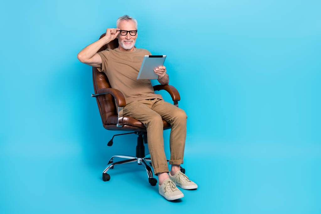 Ganzes Porträt von fröhlichen alten Mann sitzen Stuhl Hand berühren Brillen verwenden Tablette isoliert auf blauem Hintergrund. - Foto, Bild