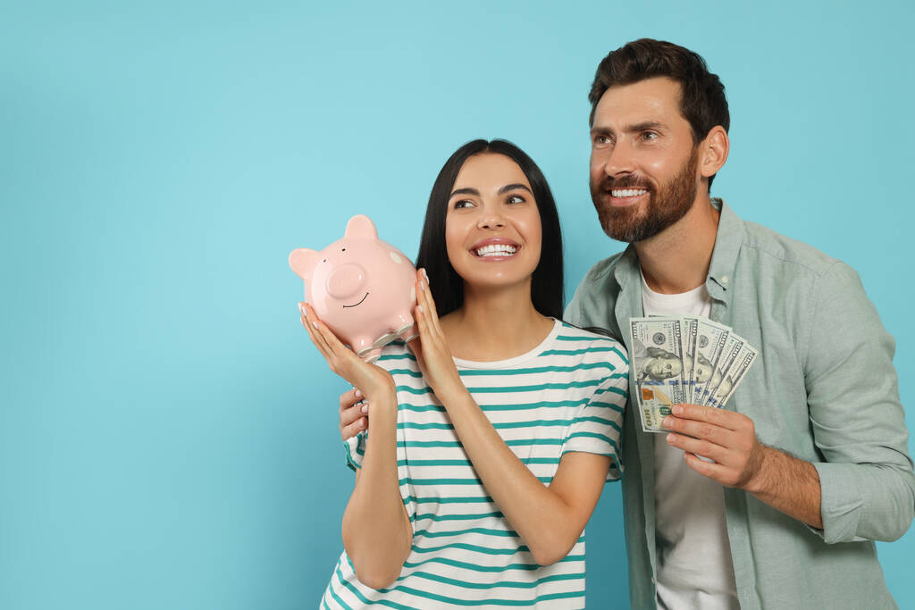 Casal feliz com dinheiro e banco piggy cerâmico no fundo azul claro, espaço para texto - Foto, Imagem