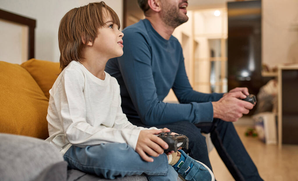 白人の少年とクロップドファーザーは自宅でソファの上でジョイスティックでビデオゲームをプレイします。集中的な子供と男は一緒に時間を過ごす。父との関係。国内レジャー・娯楽 - 写真・画像