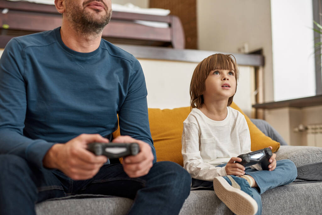 Белый мальчик и обрезанный отец играют в видеоигры с джойстиками на диване дома. Сосредоточенный ребенок и мужчина проводят время вместе. Отцовство и воспитание. Досуг и развлечения - Фото, изображение