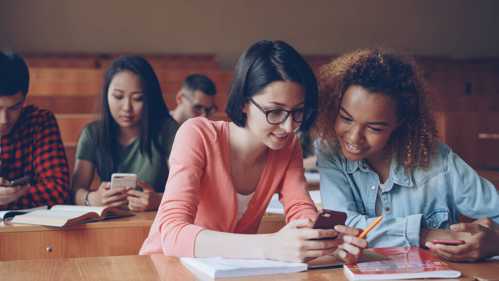 Hübsche Studentinnen nutzen Smartphone, sehen Bildschirm, reden und lachen, wenn sie an den Tischen der Universität sitzen. Soziale Medien, Internet, Millennials und Bildungskonzept. - Foto, Bild