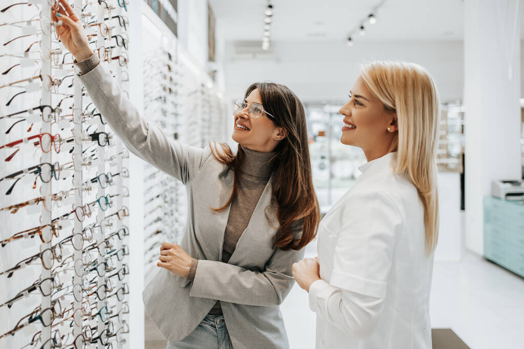 Gyönyörű és divatos nő választott szemüveg keret a modern optikai boltban. Női eladó szakember segít neki, hogy a helyes döntést. - Fotó, kép