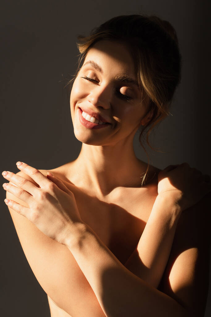 счастливая обнаженная женщина с закрытыми глазами, прикрывающая грудь скрещенными руками в солнечном свете, изолированная на сером - Фото, изображение