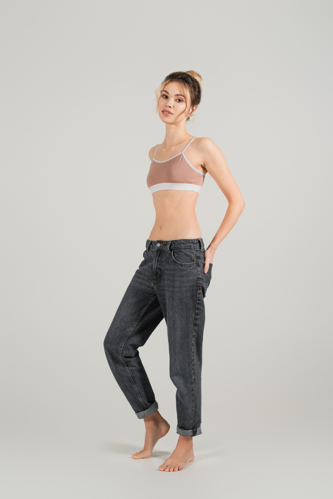 longitud completa de la mujer en forma en top deportivo y jeans de pie descalzo sobre fondo gris - Foto, Imagen